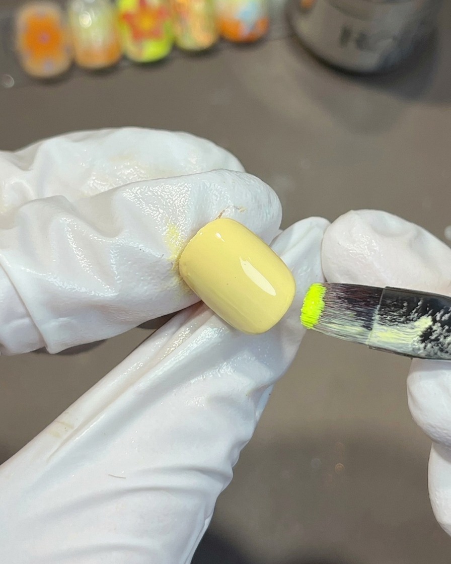 Una mano con guantes usando un pincel de uñas plano para aplicar esmalte de gel verde vibrante en la esquina de una uña pintada de amarillo.