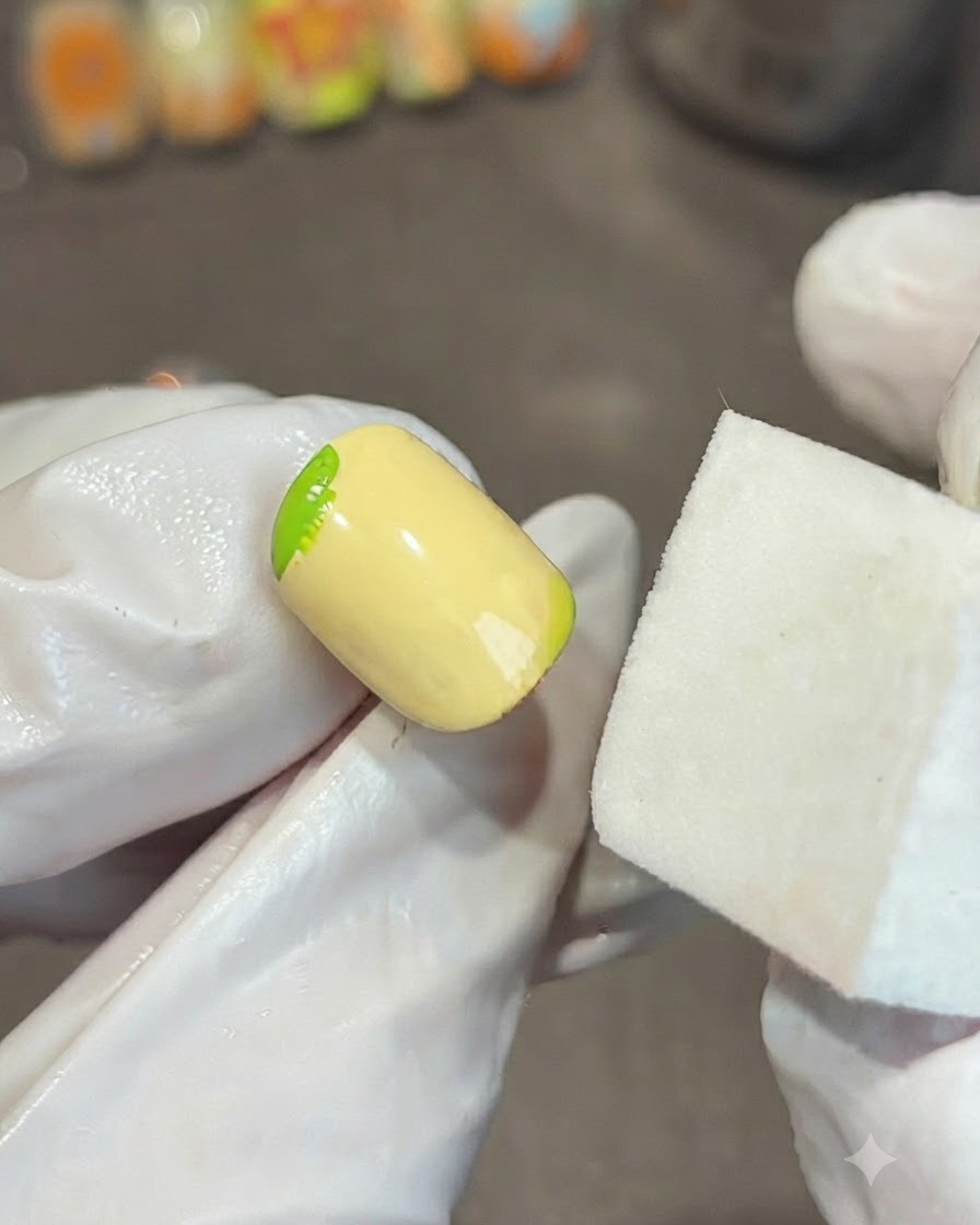 Una mano con guantes sosteniendo un bloque de esponja blanco junto a una uña amarilla, difuminando un degradado de gel verde en la esquina.