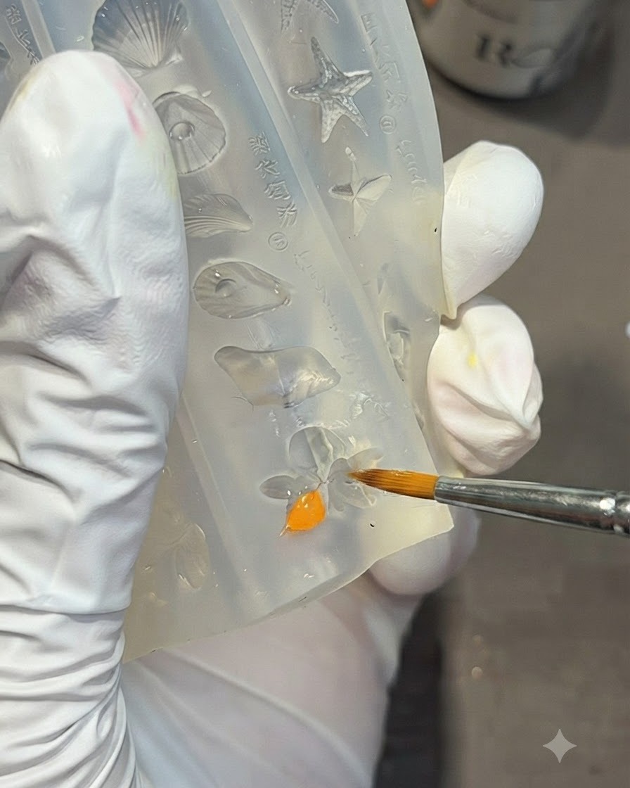 Una mano con guantes usando un pincel de detalle para rellenar la forma de una flor de lirio dentro de un molde de silicona transparente con gel naranja brillante.