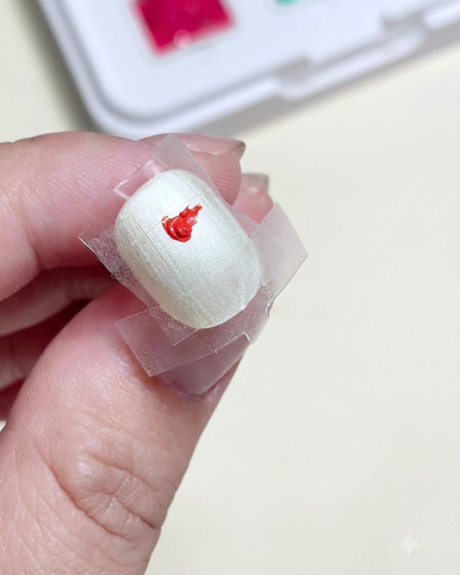 Un primer plano de una uña preparada con un cuadrado enmascarado con cinta en el centro, que muestra un punto brillante de pintura de gel roja.