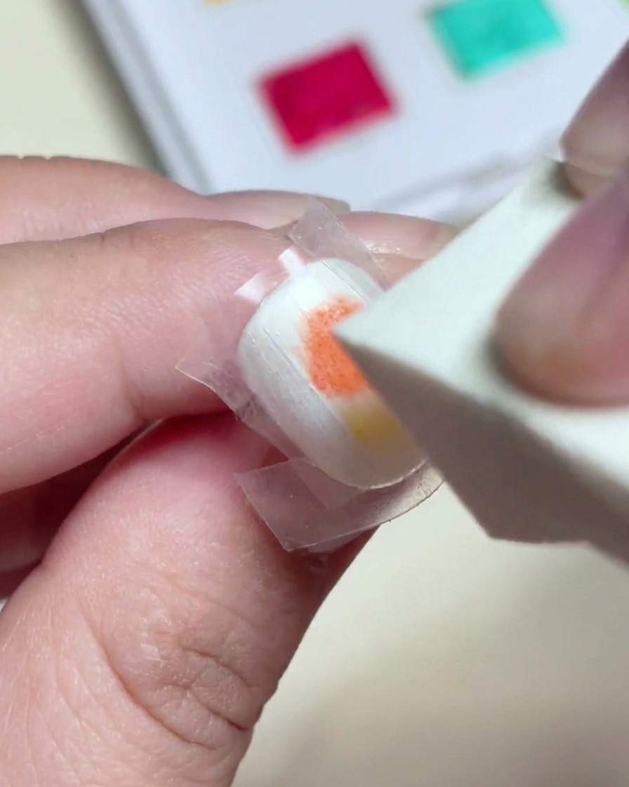 Una esponja de maquillaje dando golpecitos suaves sobre pintura de gel roja y amarilla húmeda para crear un degradado perfecto en la uña.