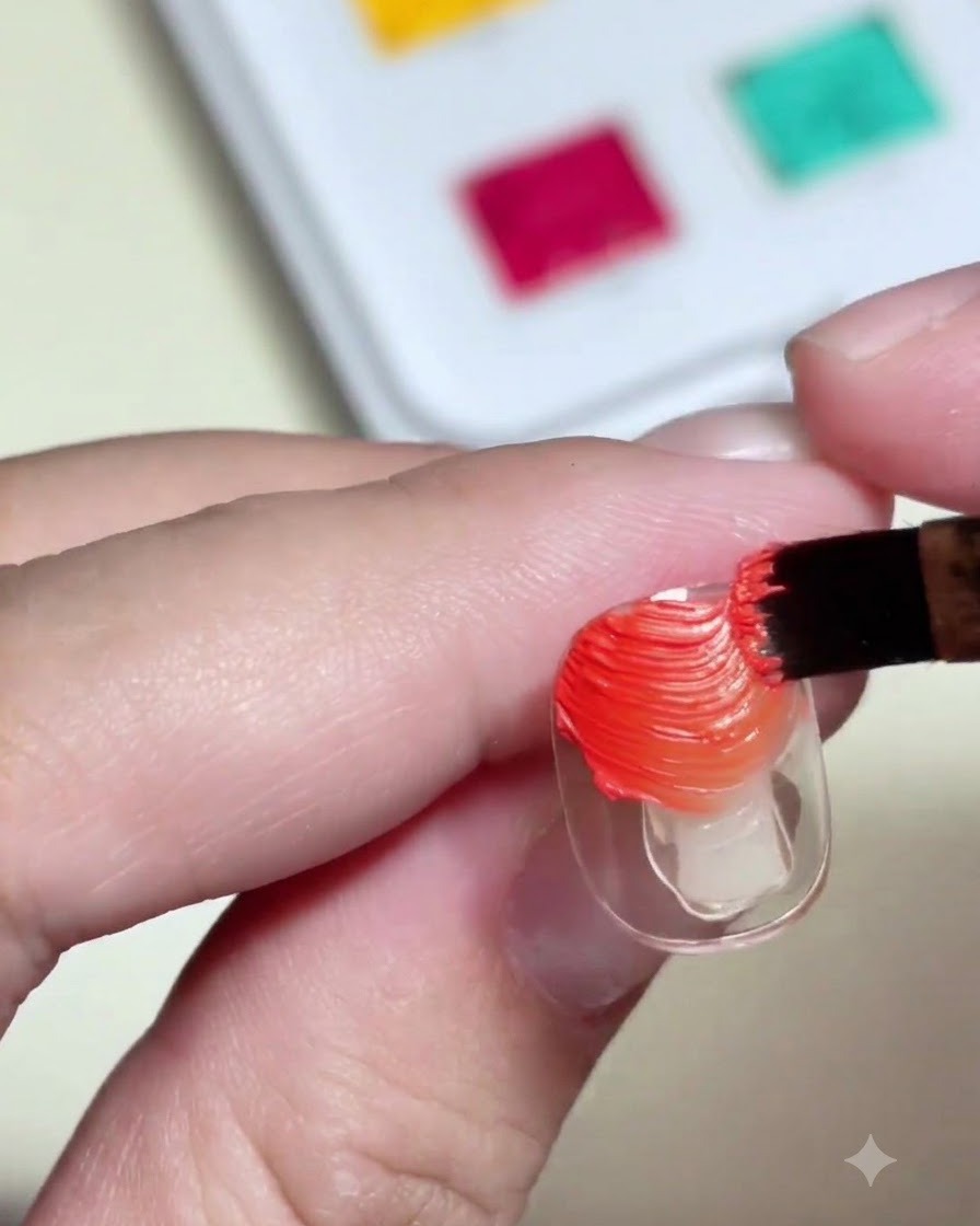 Un pincel para uñas aplicando gel de pintura de color naranja y amarillo texturizado sobre un tip de uña transparente.