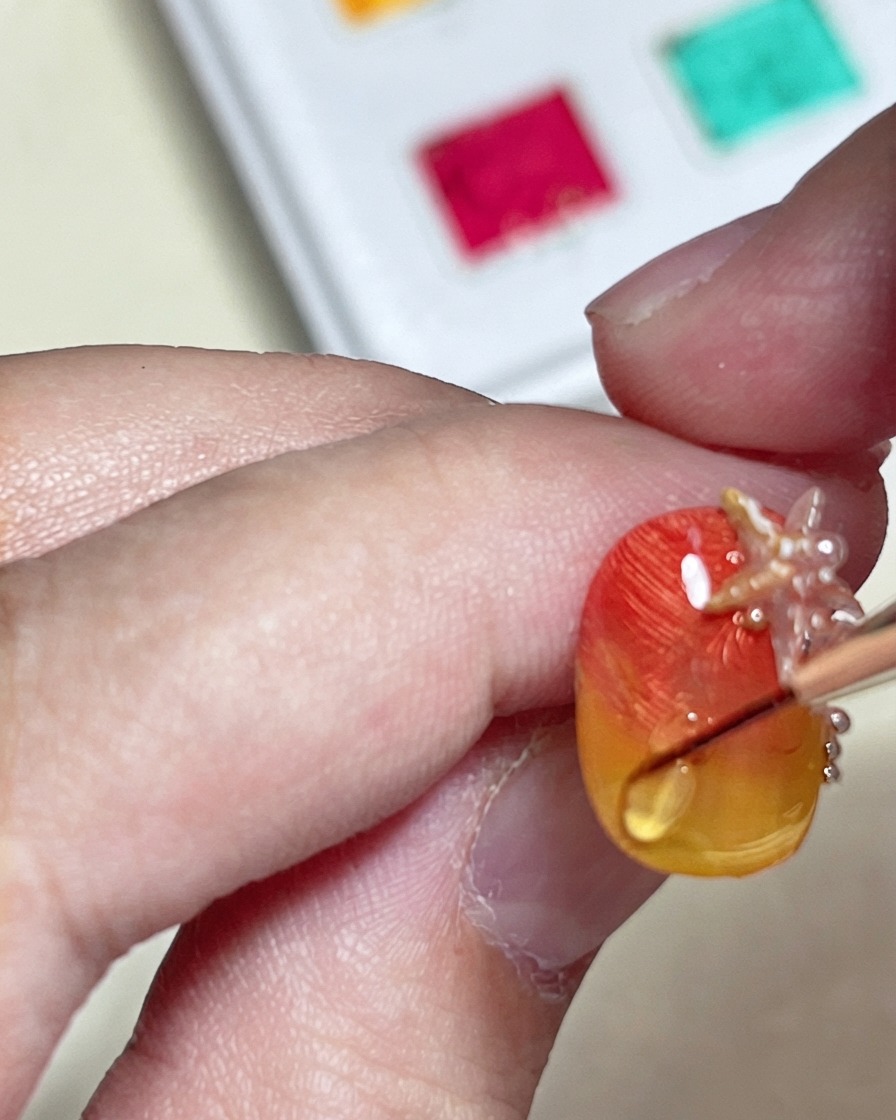 Un pincel dibujando una línea brillante de onda de agua en relieve con un top coat duro sobre una uña texturizada con degradado naranja y amarillo.