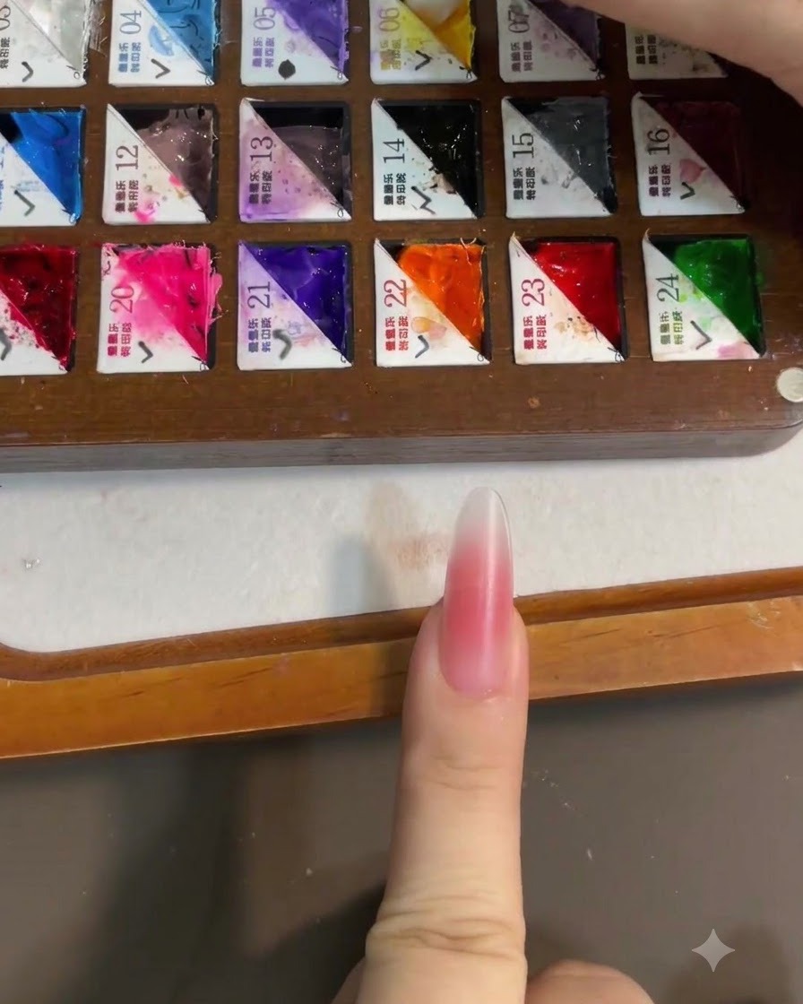 Un primer plano de un pincel tomando esmalte en gel amarillo brillante de una paleta de pintor dividida, con una uña con rubor rosa en primer plano.