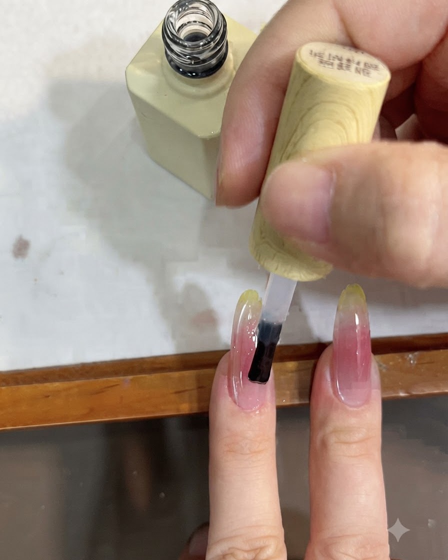 Un primer plano de un pincel aplicando una capa superior transparente sobre una uña larga con un degradado rosa y amarillo.