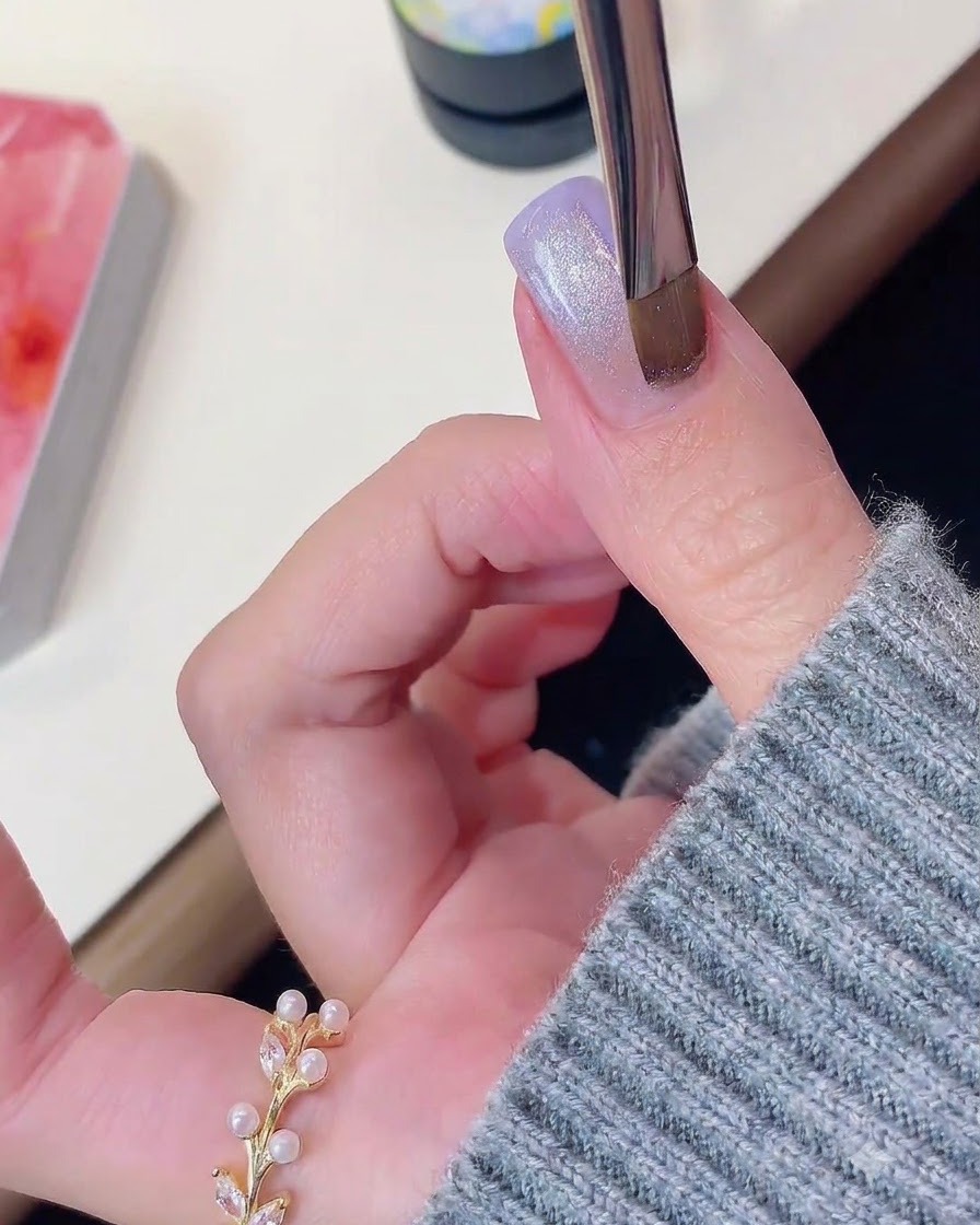 Un pincel de uñas o herramienta magnética flotando sobre una uña de gel morada brillante con efecto ojo de gato.