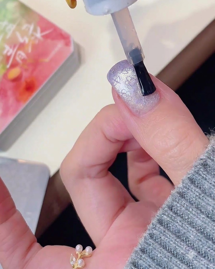 Un pincel de uñas aplicando top coat sobre un ojo de gato morado y un diseño de uñas estampado.
