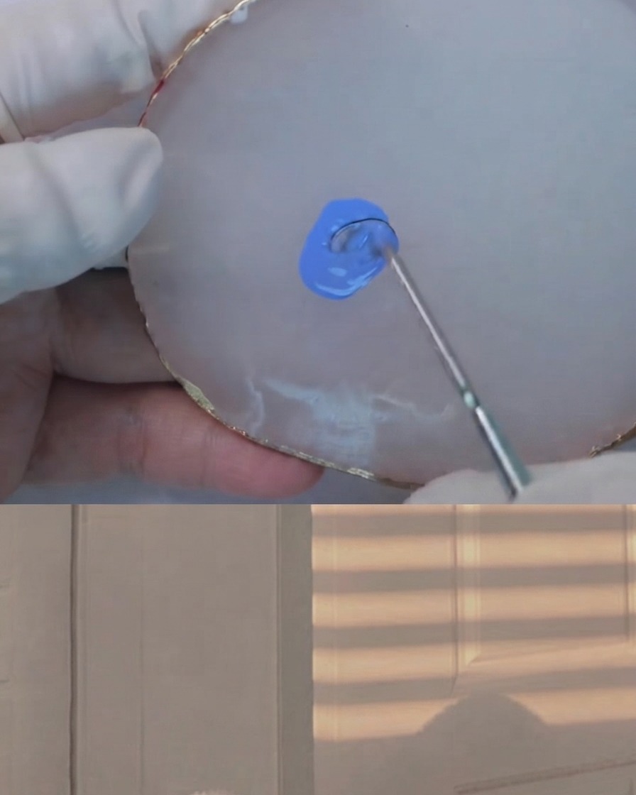 Una herramienta de mezcla incorporando una cantidad diminuta de gel oscuro en una mezcla azul claro sobre una paleta de ágata blanca.