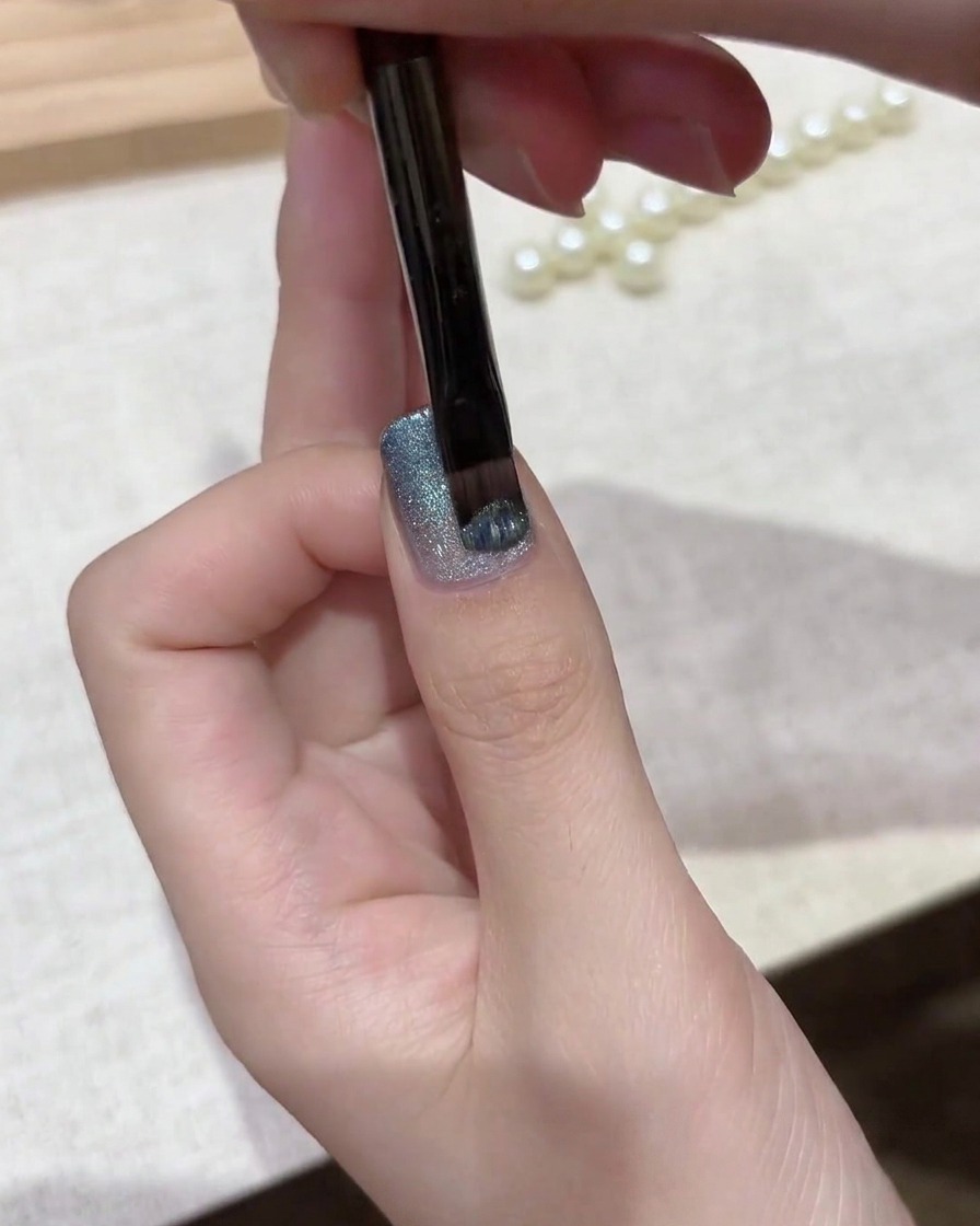 Un pincel de gel aplicando una capa de esmalte magnético brillante de ojo de gato azul plateado sobre una base degradada.