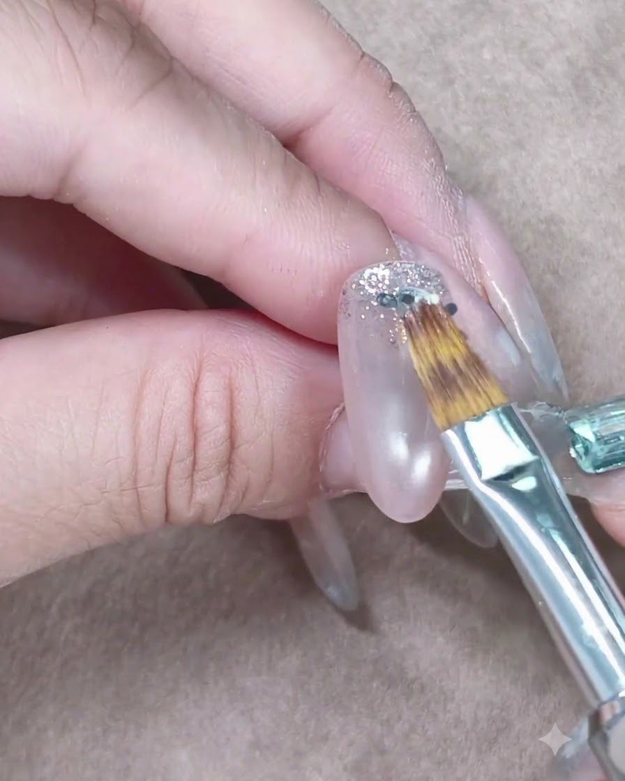 Un pincel de uñas pequeño esparciendo suavemente la brillantina plateada hacia abajo en una uña de gel transparente.