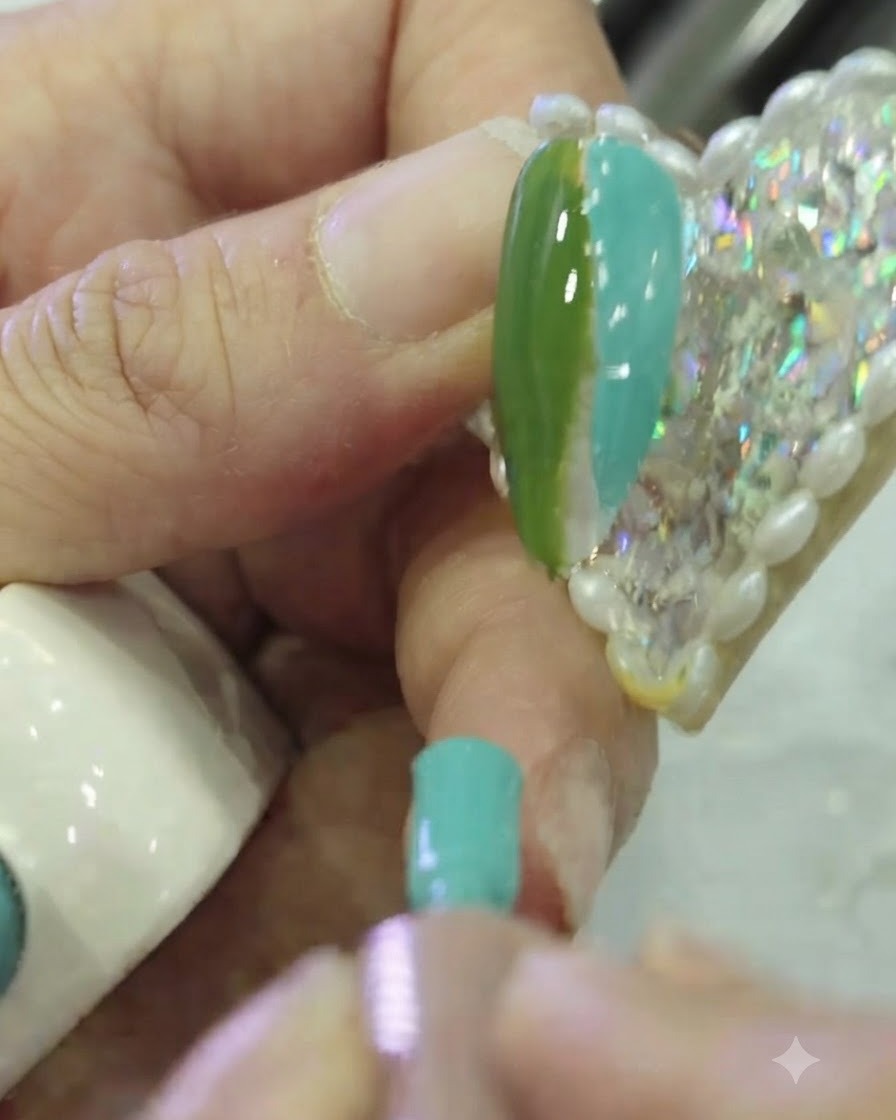 Primer plano de un pincel plano para nail art dando ligeros toques en la frontera entre el esmalte en gel verde oscuro y turquesa sobre la uña.