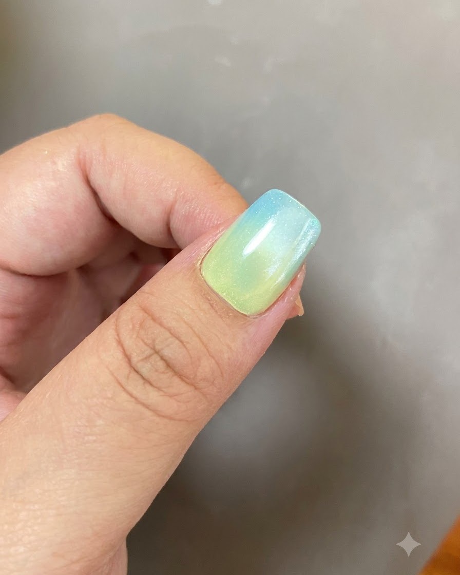 Una manicura de gel completada con un degradado de azul y amarillo y un diseño de anillo brillante bajo iluminación suave.