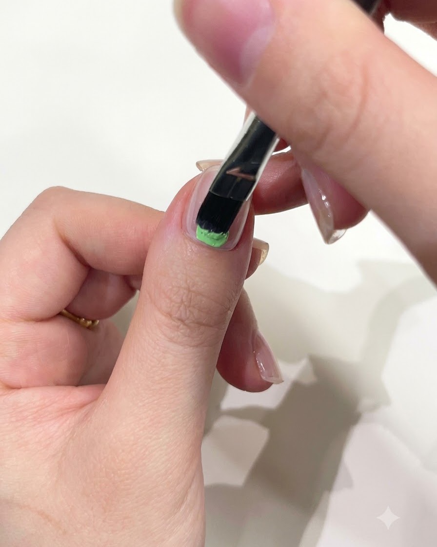 Un primer plano de una mano utilizando un pincel plano para gel para aplicar gel sólido verde menta en la base de la uña del pulgar.