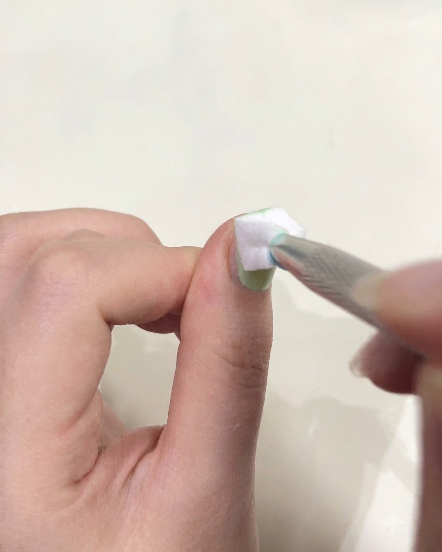 Un primer plano de una mano sosteniendo unas pinzas con una pequeña esponja blanca aplicando golpecitos de gel verde sobre una uña.