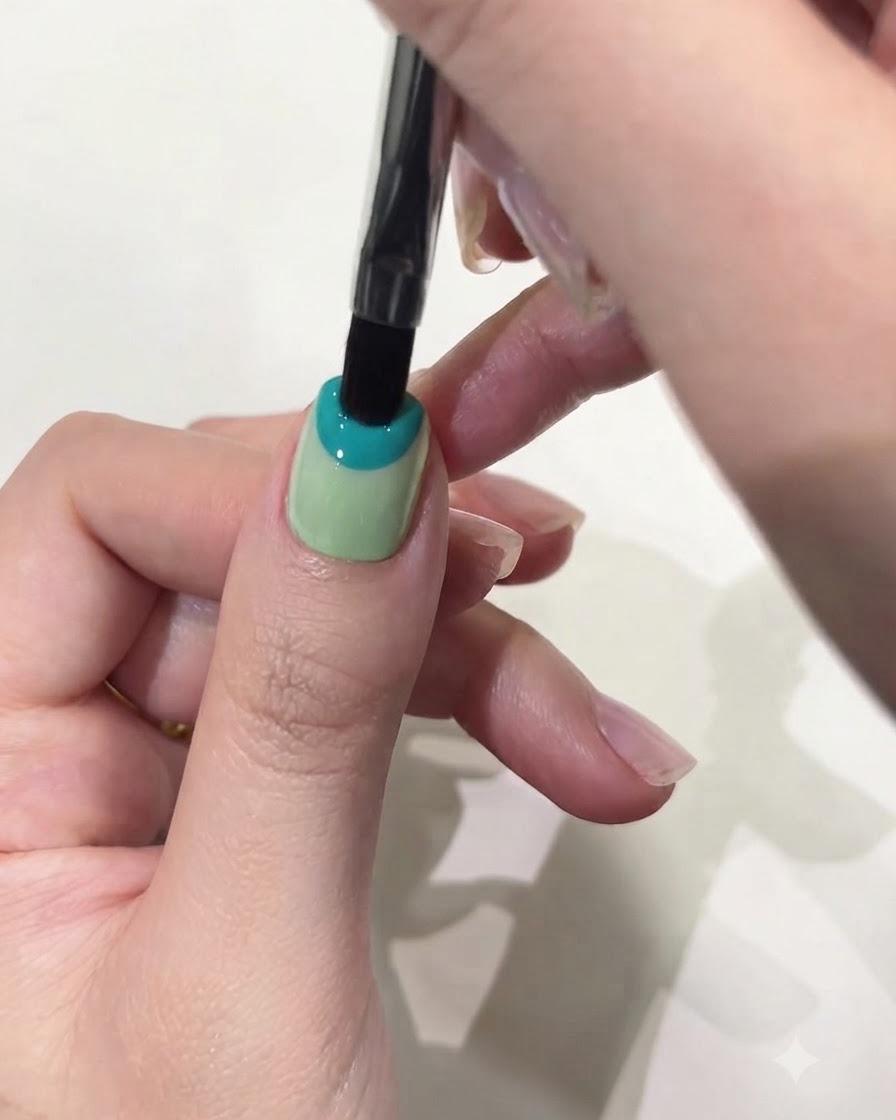 Un primer plano de un pincel plano aplicando gel sólido azul pastel en la mitad superior de la uña del pulgar.