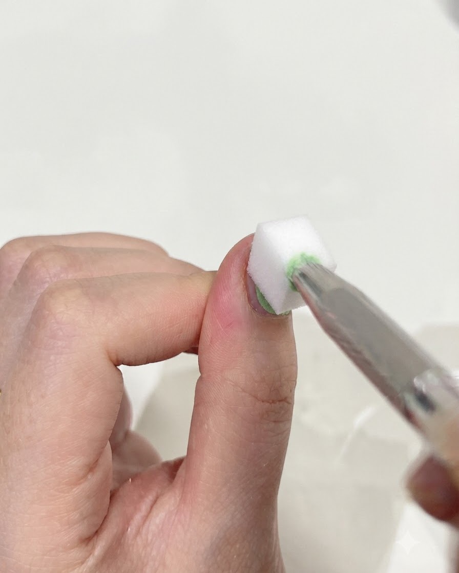 Un primer plano de unas pinzas sosteniendo una pequeña esponja de maquillaje blanca, dando golpecitos suaves en la superficie de una uña para mezclar los geles verde y azul.