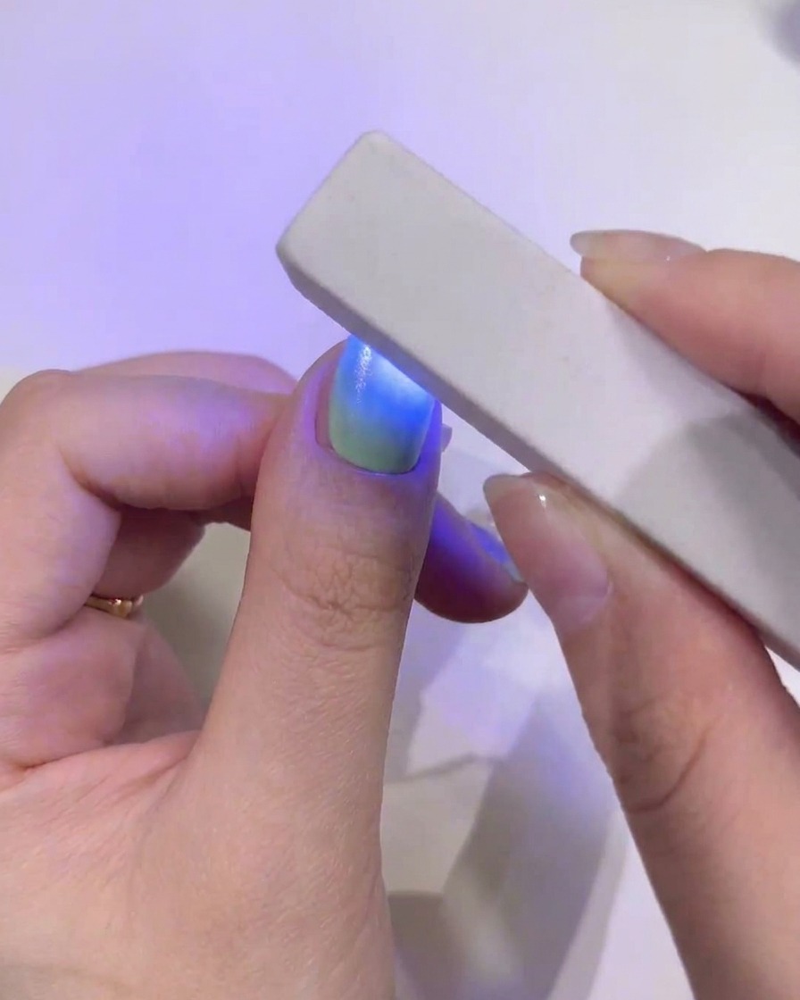 Un primer plano de la uña del pulgar con un degradado continuo en verde y azul siendo curada bajo una mini lámpara UV/LED azul de mano.