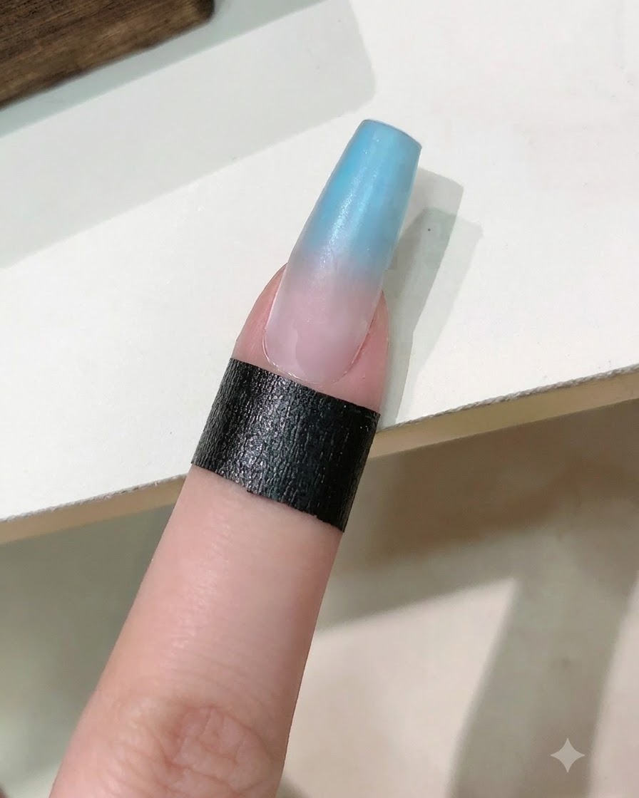 Un pincel de uñas aplicando una capa transparente de gel reafirmante sobre la uña con degradado azul y rosa.