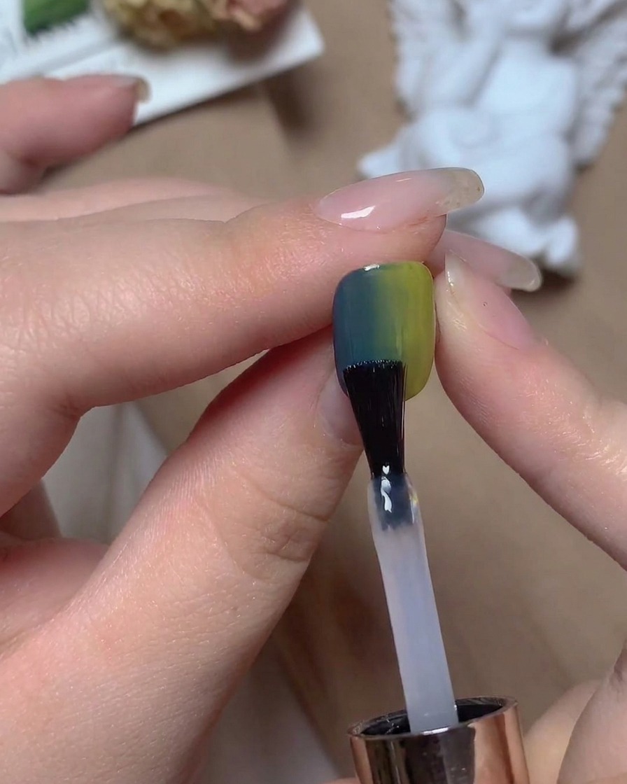 Un pincel de uñas aplicando un top coat transparente sobre una uña degradada azul y verde bajo cálida iluminación de estudio.