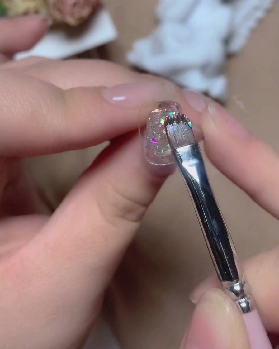 Un pincel fino aplicando gel con purpurina multicolor brillante sobre la superficie de una uña con reflejos vibrantes.
