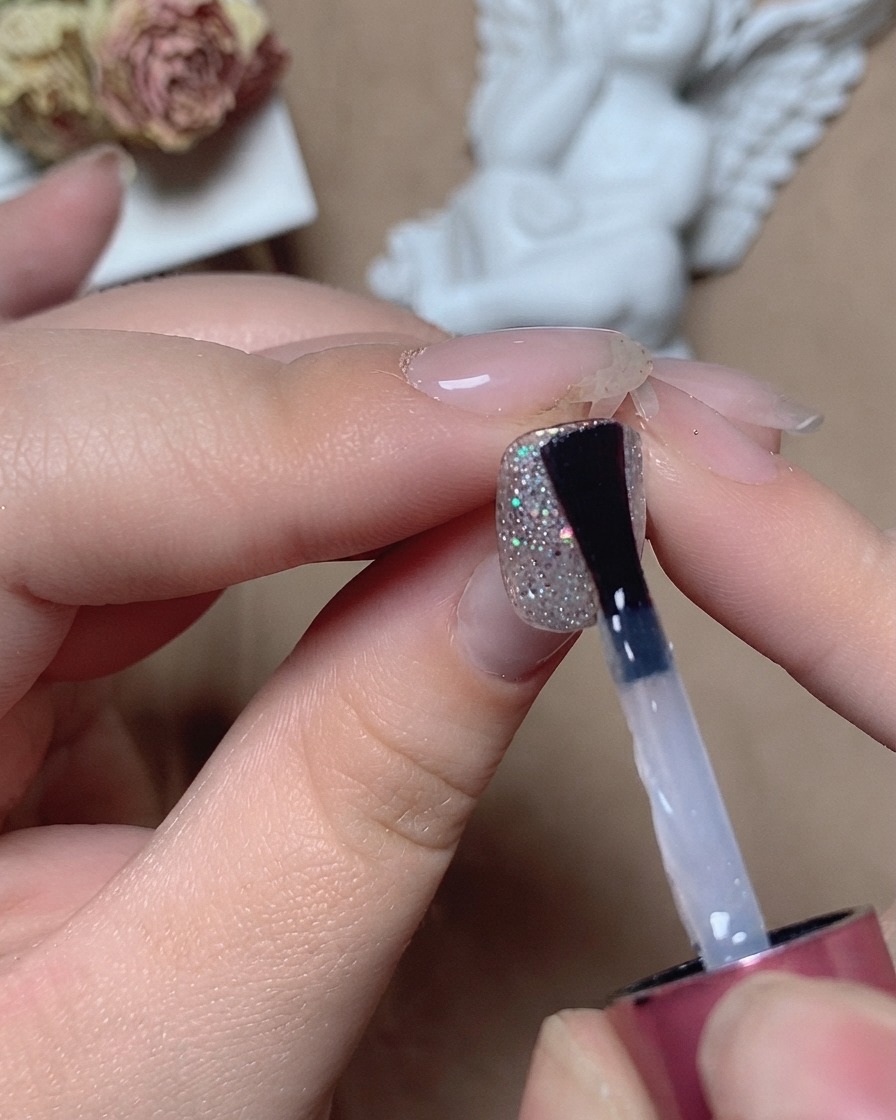 Un pincel aplicando un top coat nivelador transparente sobre una uña texturizada con purpurina bajo iluminación de estudio.