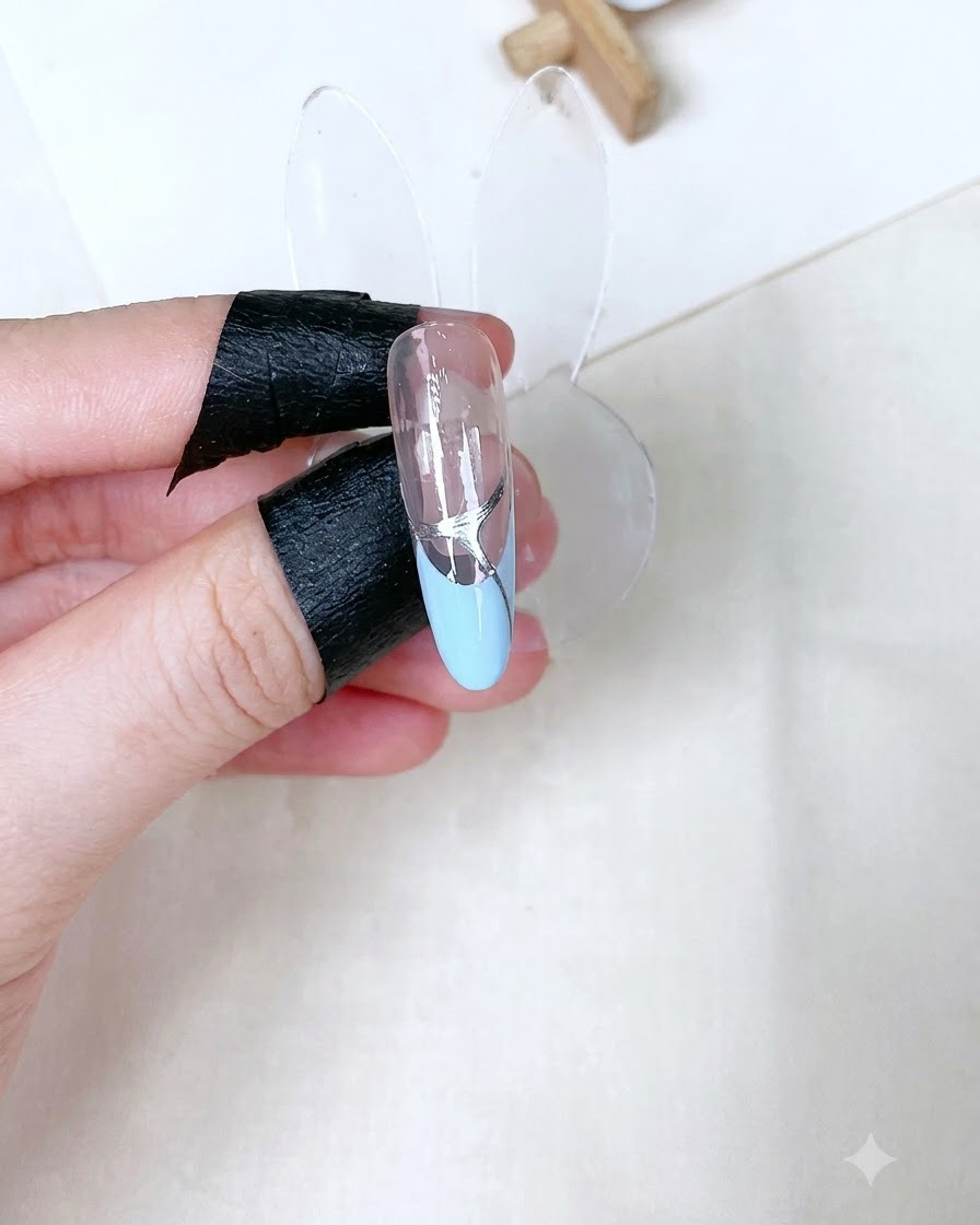 Un soporte de uña esculpida individual que muestra una capa superior transparente brillante curada con detalles plateados sobre una punta francesa azul bebé.