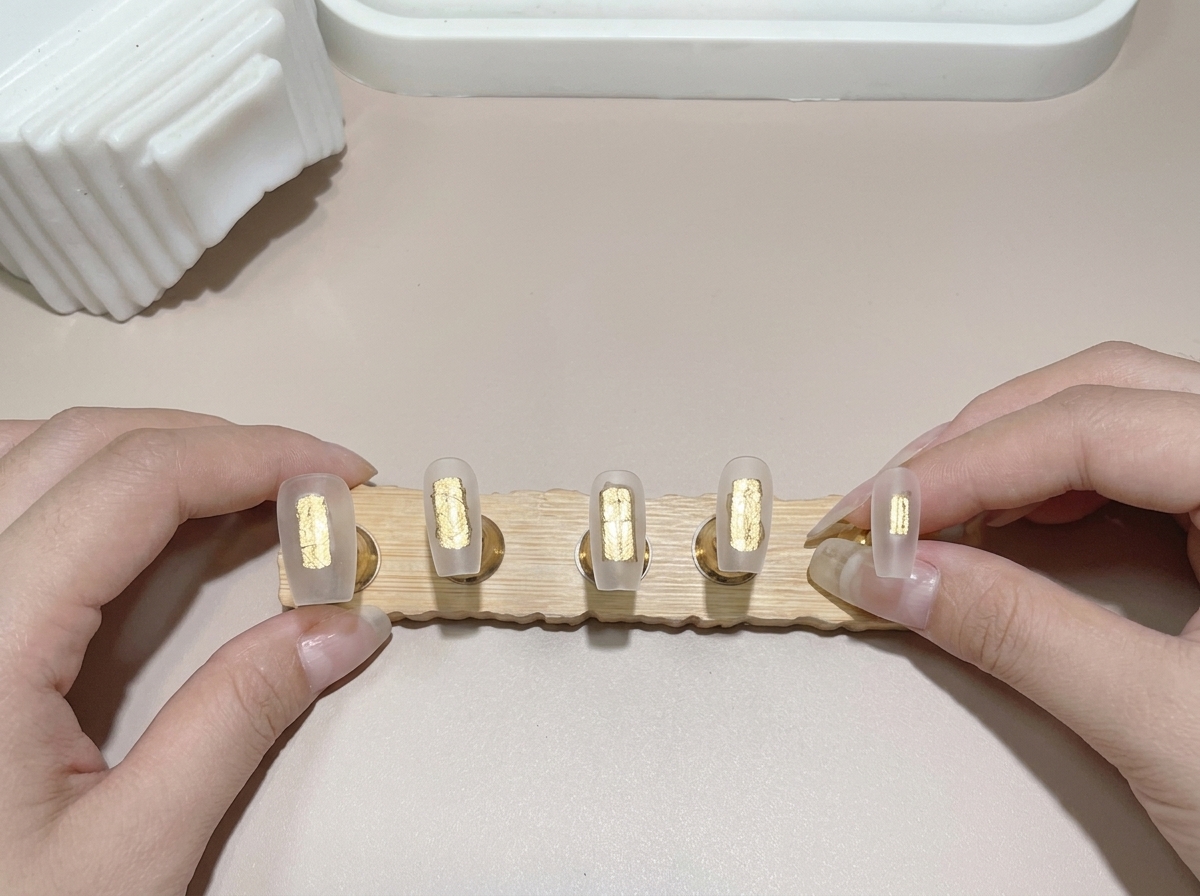 Un primer plano de un pincel aplicando gel base en múltiples puntas de uñas montadas en un soporte de madera.