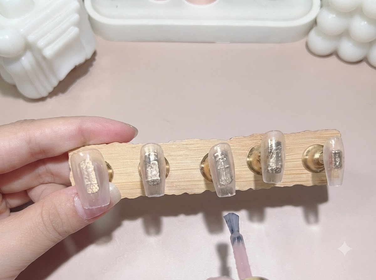 Un pincel de esmalte en gel aplicando una segunda capa de color rosa lechoso traslúcido a las puntas de las uñas adheridas a un soporte de exhibición de madera.