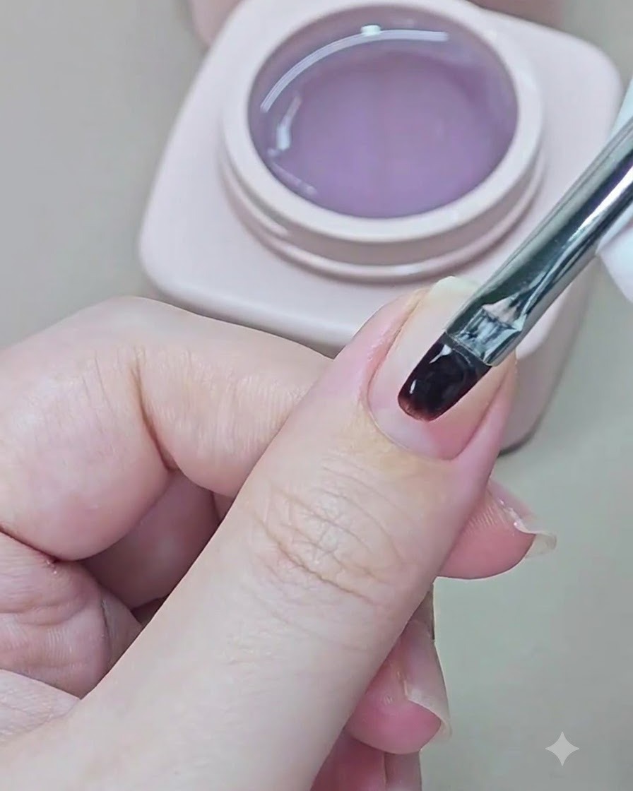 Un pequeño pincel de uñas aplicando gel base transparente sobre una uña natural.