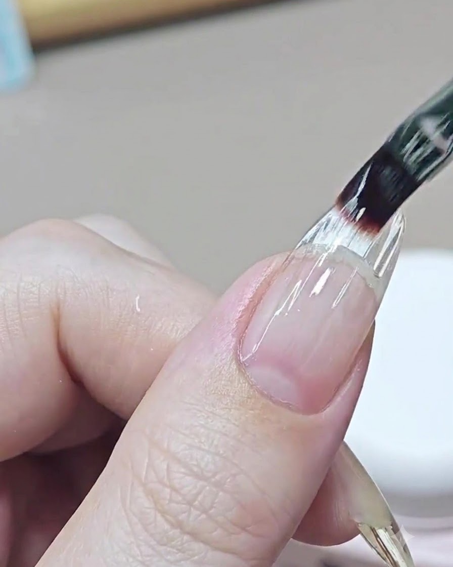 Un pincel fino de uñas aplicando una capa transparente y niveladora de gel constructor sobre el área de transición de una uña extendida.