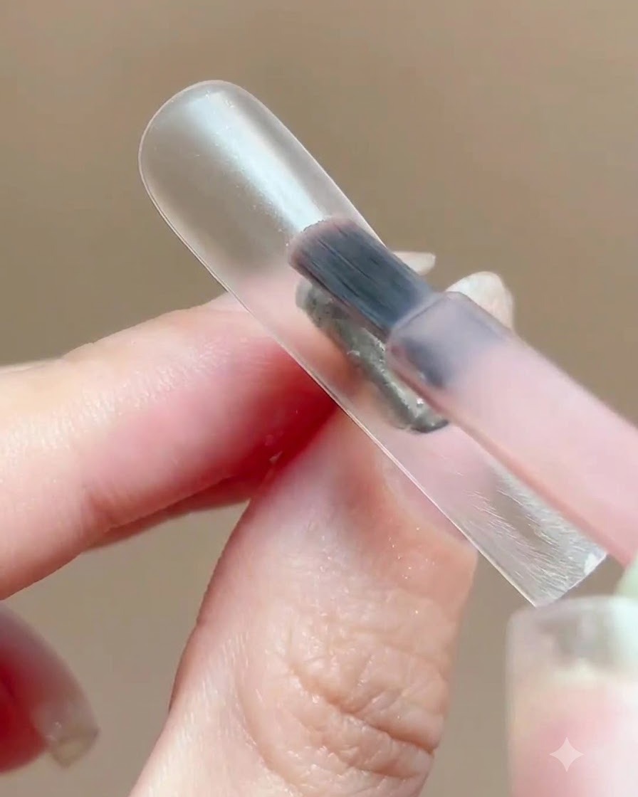 Un pincel aplicando una capa fina de esmalte en gel rosa translúcido sobre un soporte transparente para exhibir uñas.