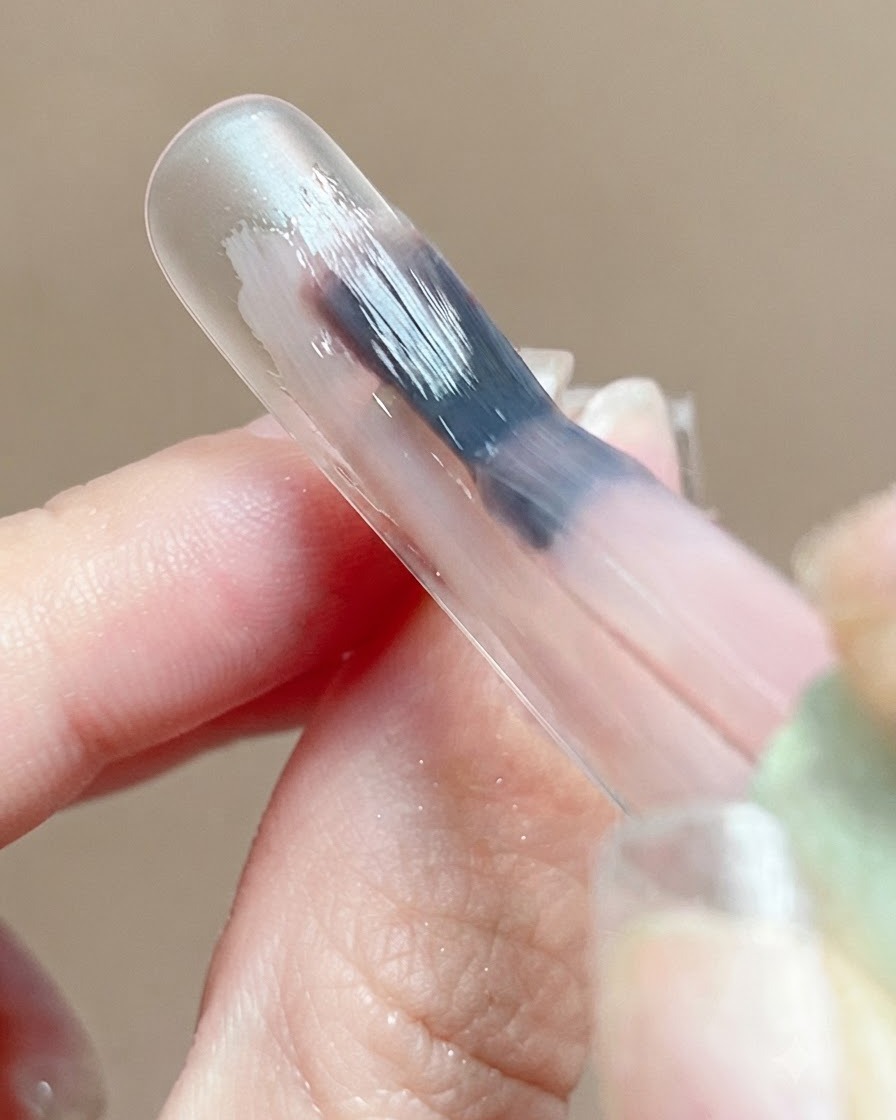 Un primer plano de una capa base de gel rosa reflejando la luz suavemente en la superficie de una uña durante la autonivelación.