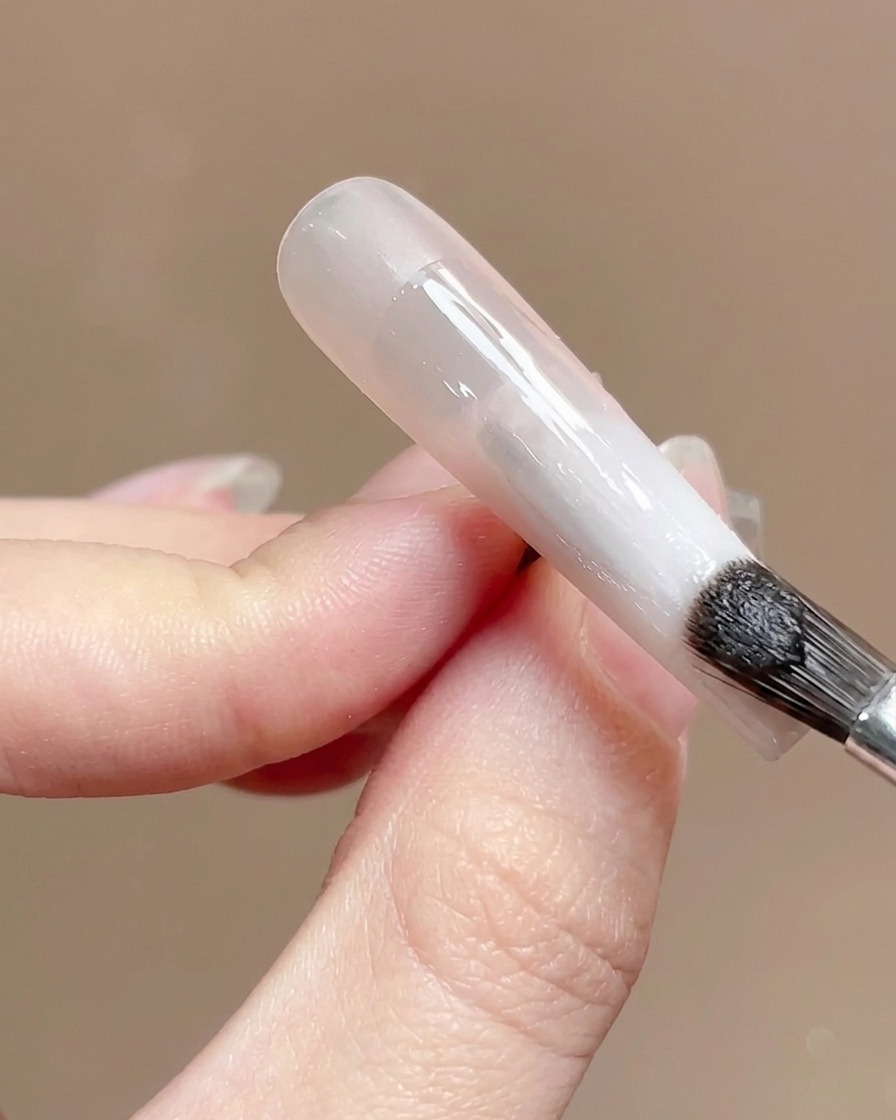 Un pincel de uñas aplicando un top coat transparente y brillante sobre la superficie de una uña con degradado.