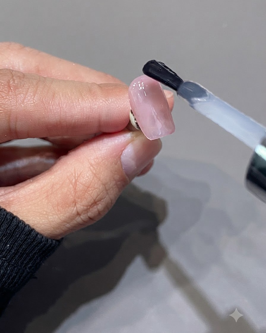 Un pincel aplicando una capa superior rosa lechosa translúcida sobre una superficie de uña rosa brillante.