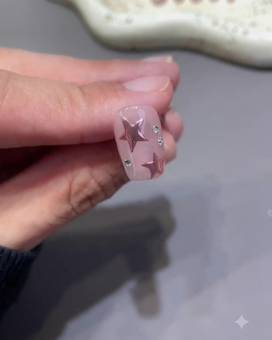 Un pincel de detalle diminuto pintando meticulosamente la capa superior alrededor de la base de pequeños strass en una uña terminada con estrellas rosas.