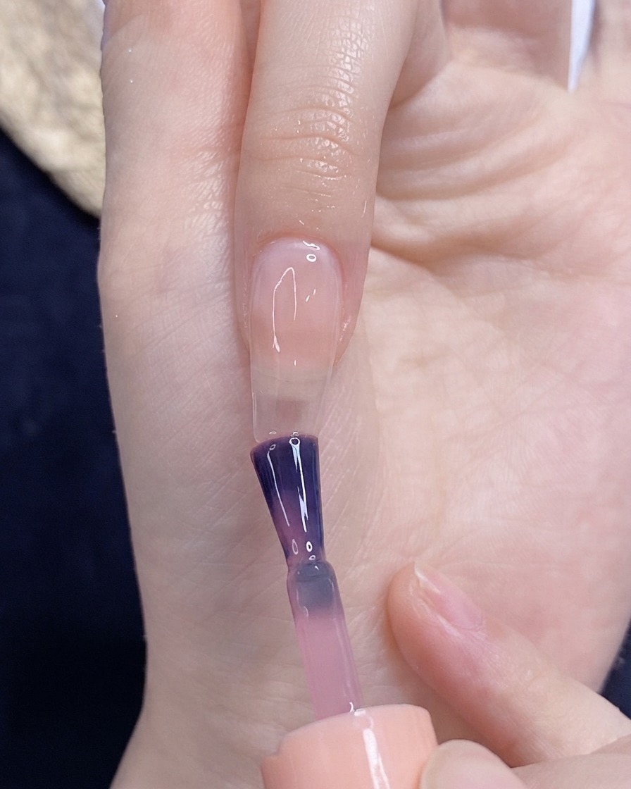 Un pincel aplicando esmalte de gel rosa traslúcido sobre una uña natural.