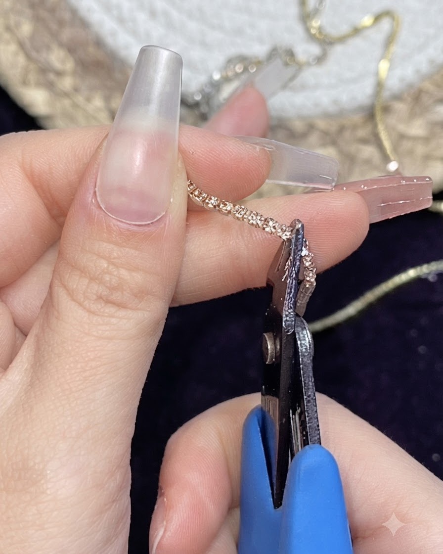 Un par de alicates pequeños de metal cortando una tira de cadena metálica dorada sostenida entre los dedos.