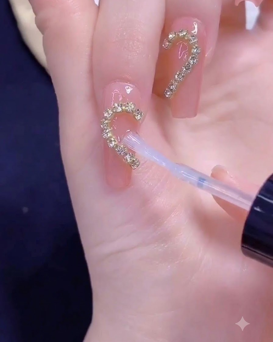 Un pincel aplicando top coat transparente sobre una tira de cadena dorada dispuesta en forma de medio corazón en una uña.