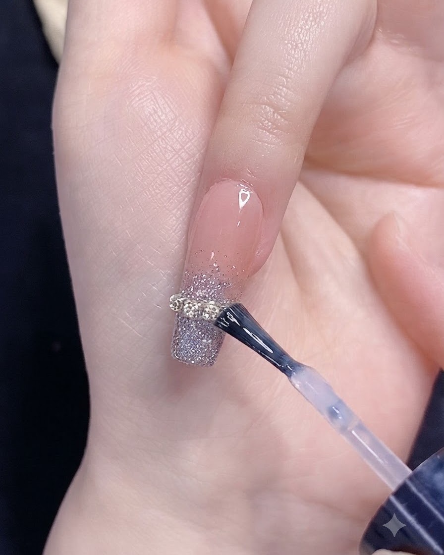 Un pincel aplicando top coat sobre una uña con degradado de purpurina plateada y un adorno de tira de cadena.