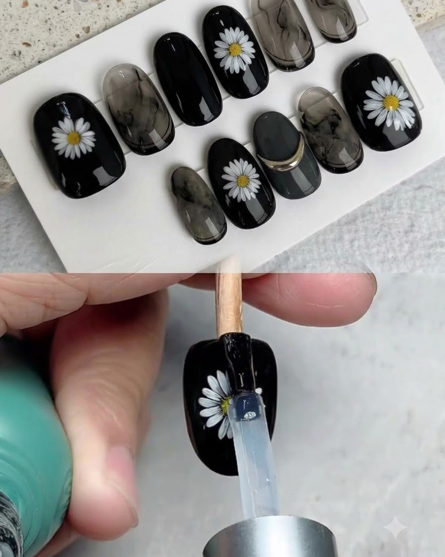Un pincel aplicando una capa superior brillante sobre una uña postiza negra terminada y decorada con una margarita blanca.