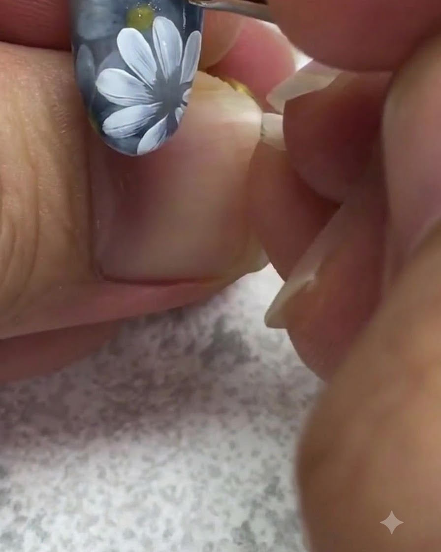 Un pincel de detalle fino aplicando un punto brillante de gel amarillo en el centro de una margarita blanca pintada sobre una uña gris.