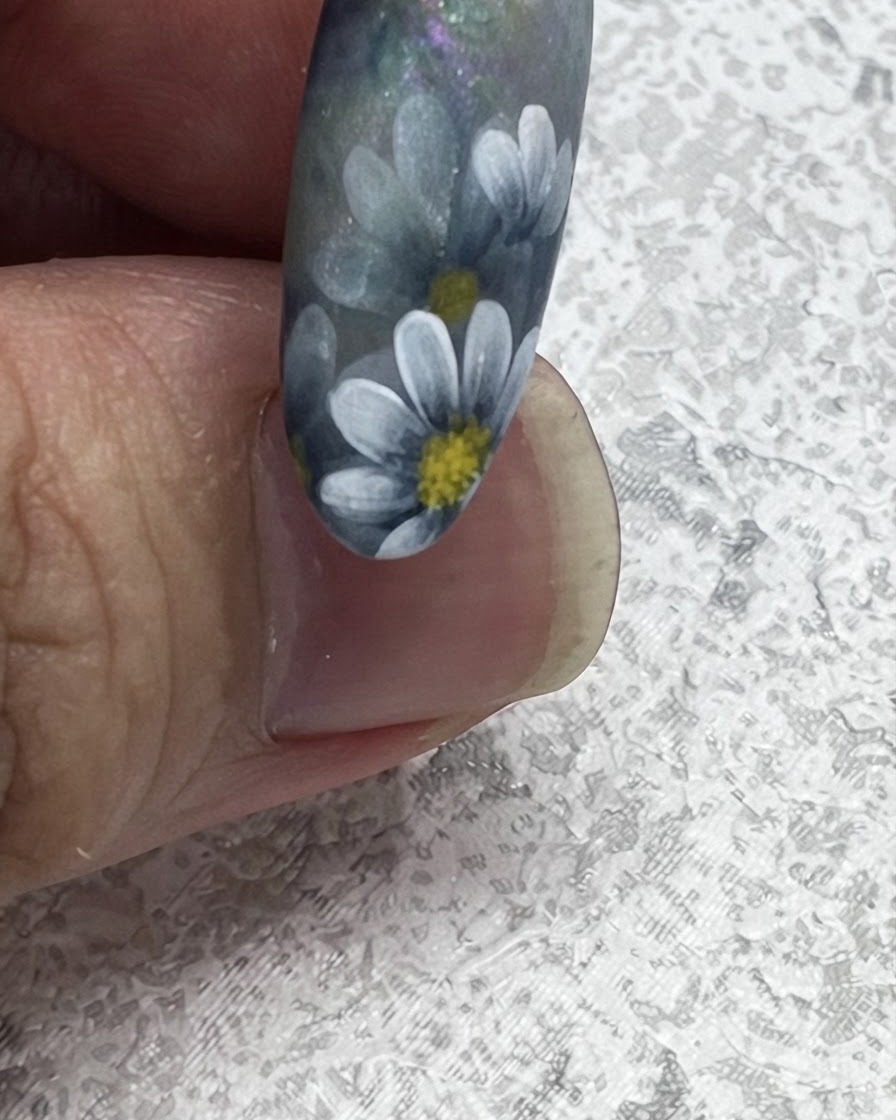 Primer plano de un pincel de uñas tirando hacia afuera para crear una punta de hoja afilada junto a las margaritas estilo nórdico pintadas.