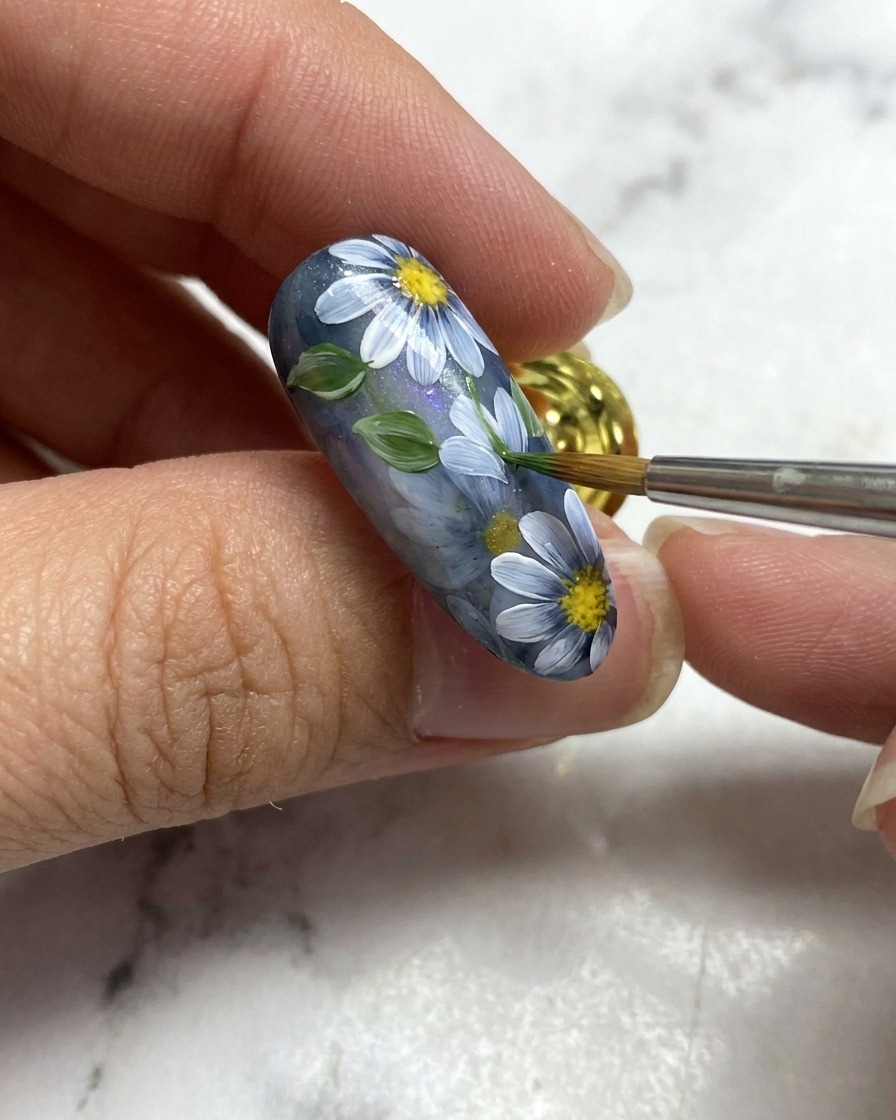 Un pincel de arte en uñas añadiendo una pequeña hoja verde junto a los detalles florales blancos sobre una base de uña gris.