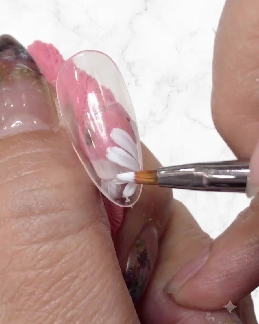 Un primer plano de un artista de uñas añadiendo más pétalos blancos al diseño de la margarita con un pincel fino en una punta de uña transparente.