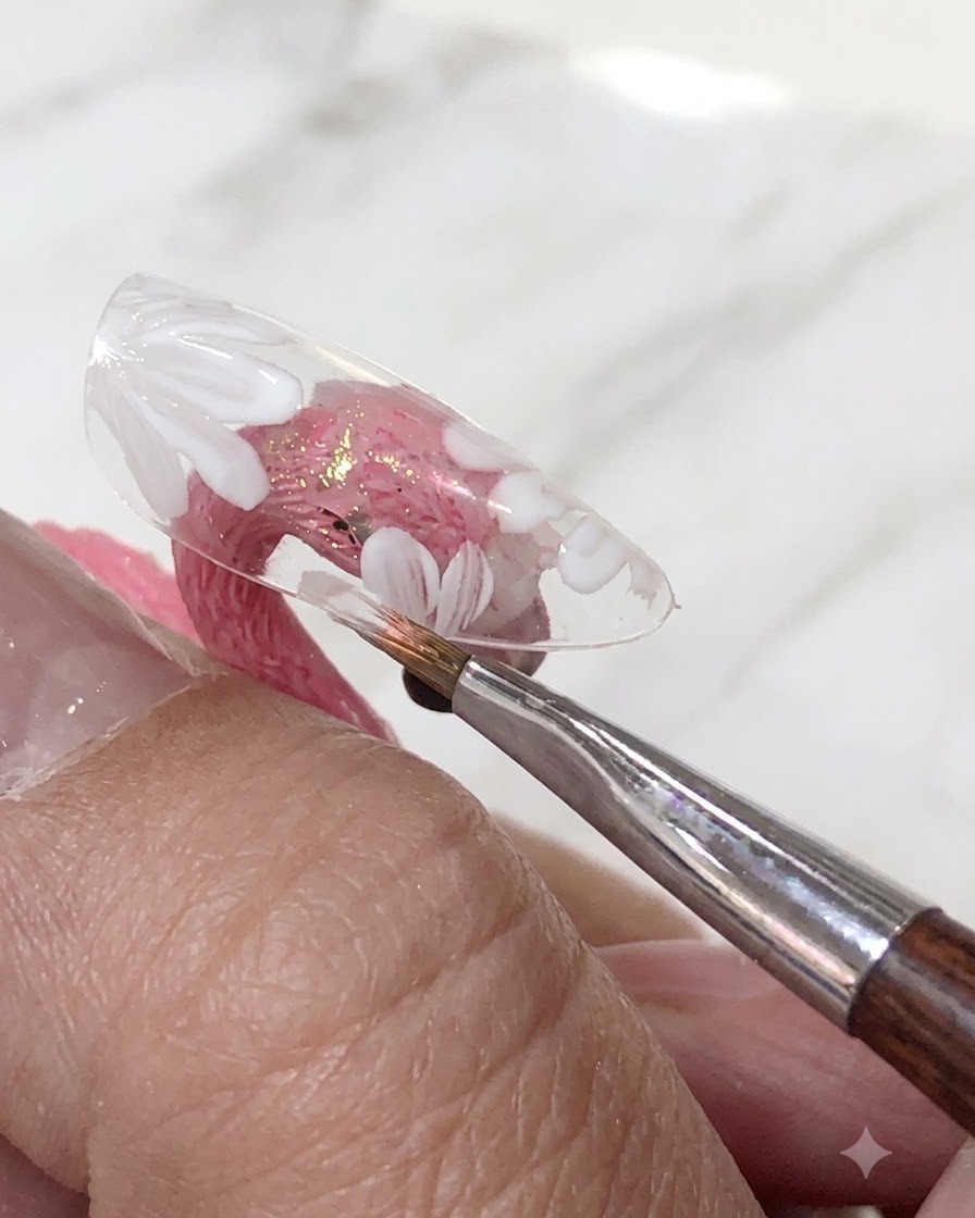 Un pincel aplicando una capa de esmalte en gel rosa translúcido sobre las flores blancas pintadas en una punta de uña transparente.
