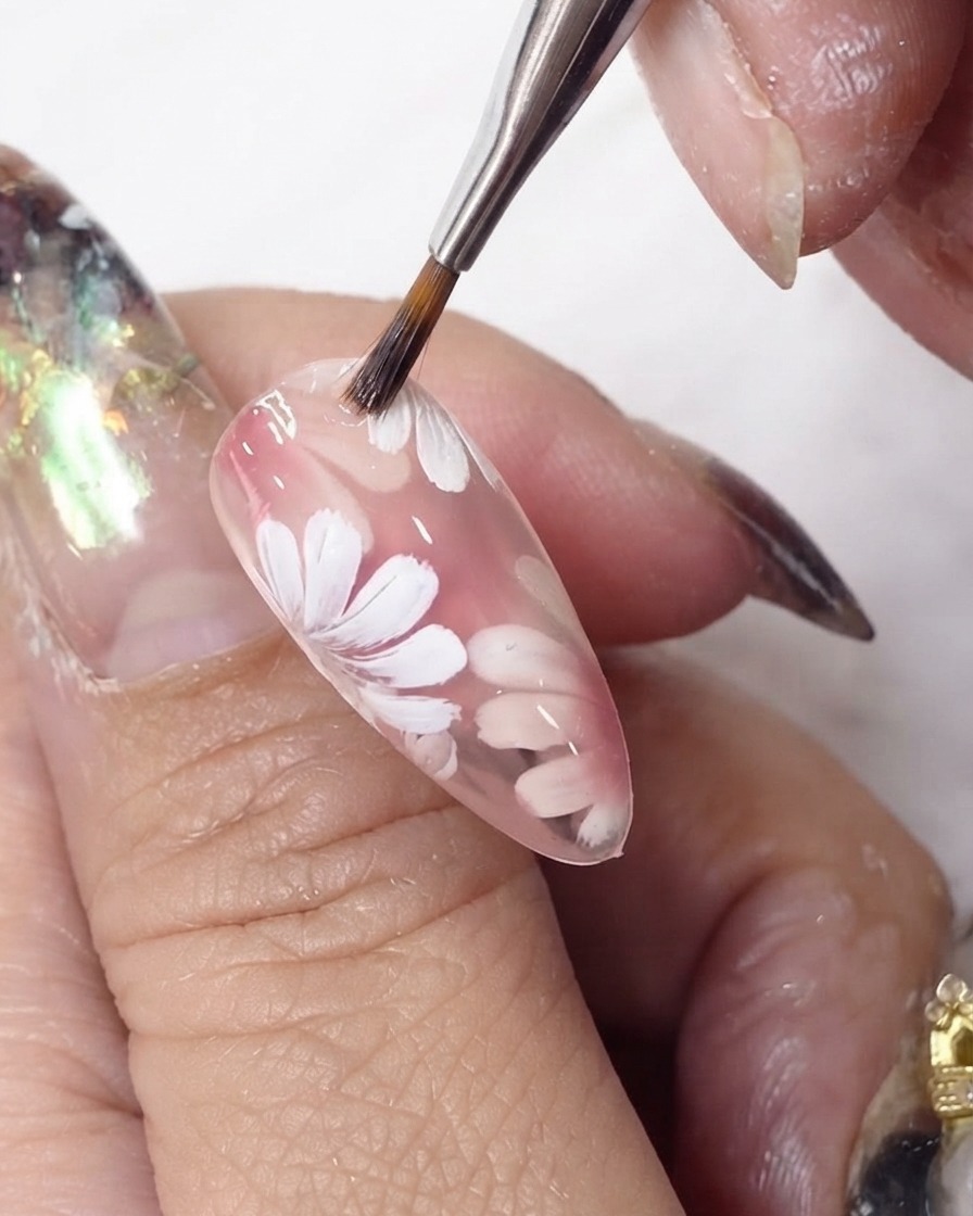 Un pincel de gel deslizando una capa de esmalte rosa transparente sobre flores blancas pintadas en una uña en forma de almendra.