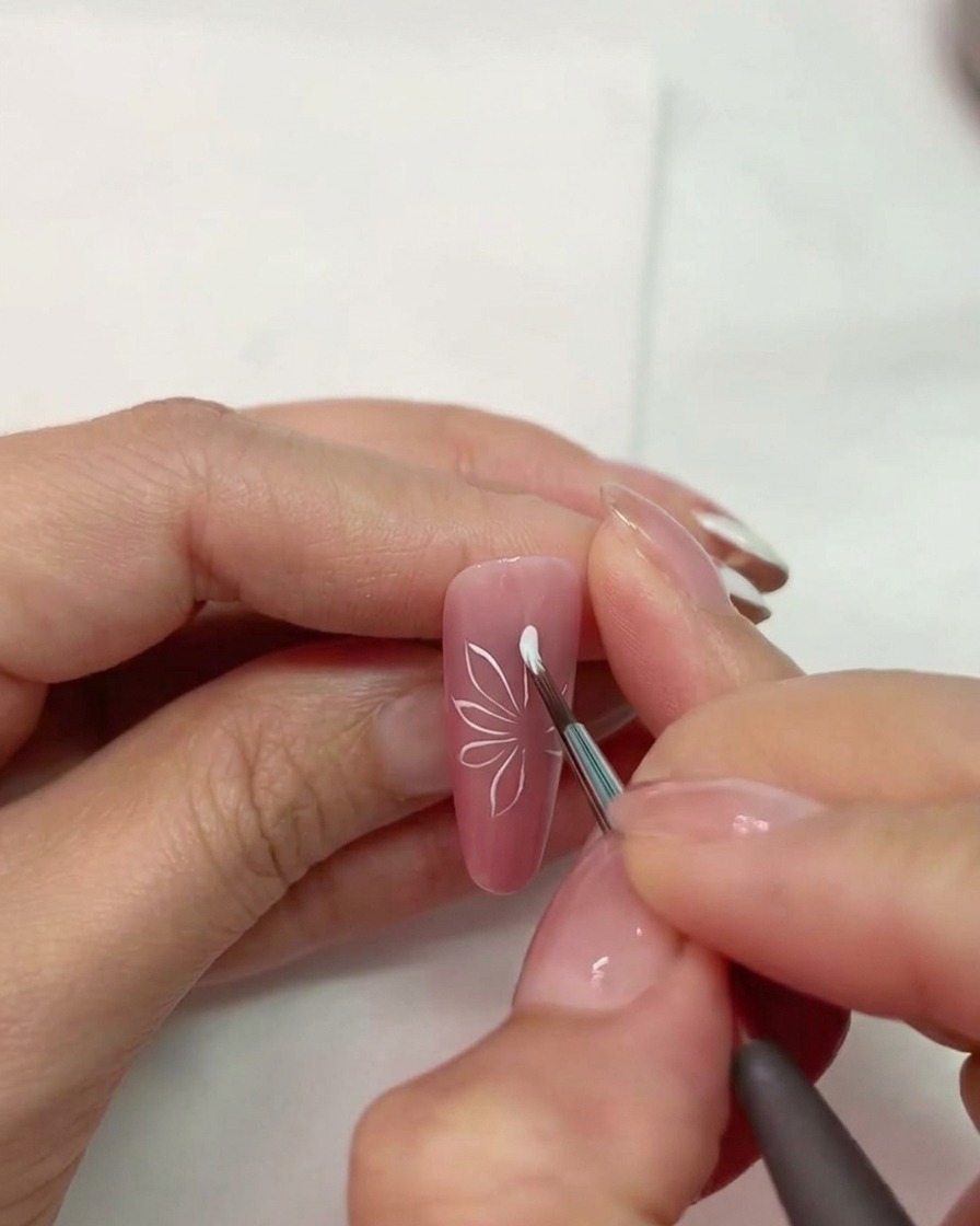 Un primer plano de un pincel de uñas dibujando formas de pétalos blancos sobre una uña rosa mate.