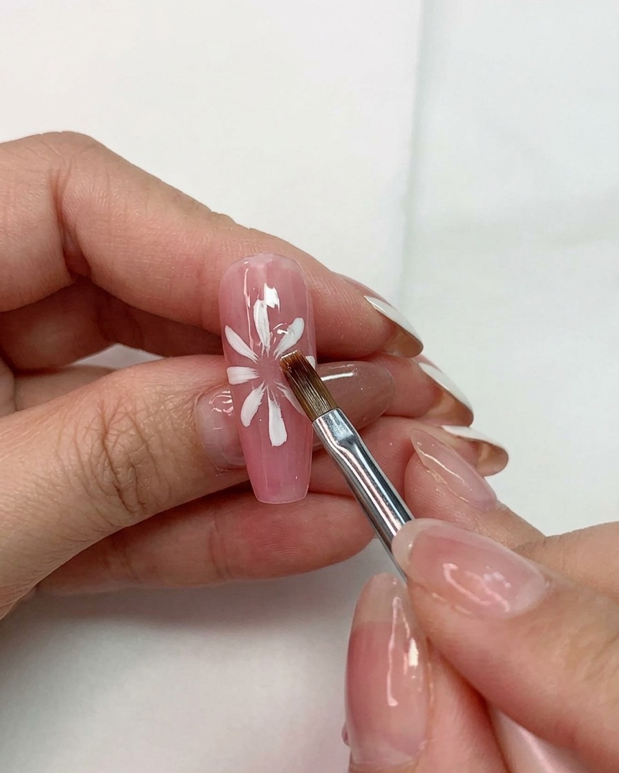 Una mano sosteniendo un pincel multiusos moldeando pétalos blancos sobre una base rosa en una uña.