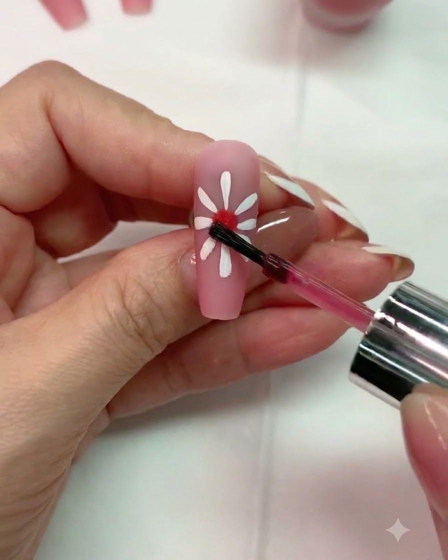 Un pincel aplicando una pequeña gota de gel blooming rojo en el centro de un diseño de margarita blanca sobre una uña rosa mate.