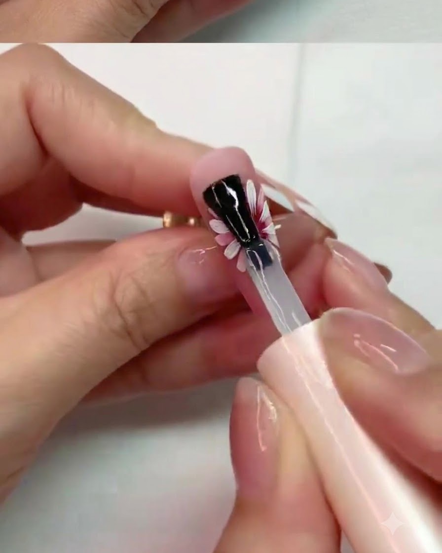 Un pincel aplicando cuidadosamente top coat mate en los pétalos del medio de un diseño de uña de margarita pintada.